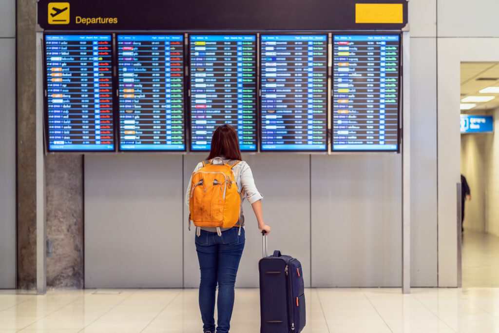 Parte traseira do viajante com bagagem em pé sobre a placa de voo para check-in na tela de informações de voo em um aeroporto moderno, viagens e transporte com conceito de tecnologia. Ela sabe sobre as regras de turismo e viagens para 2022.
