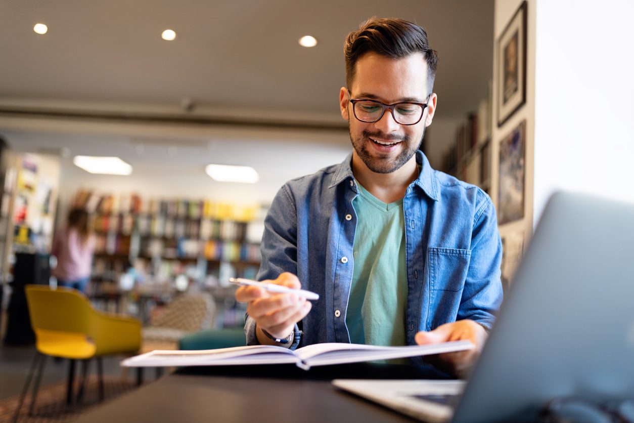 Um homem branco, de óculos preto, camisa azul e cabelo castanho está em uma biblioteca usando o computador para pesquisar os benefícios de se contratar um buscador de passagens