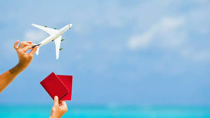 Uma mão está segurando um avião branco em miniatura e a outra mão está segurando dois passaportes vermelhos. Essa pessoa está aproveitando a alta das milhas aéreas para marcar viagens com uma agência de turismo