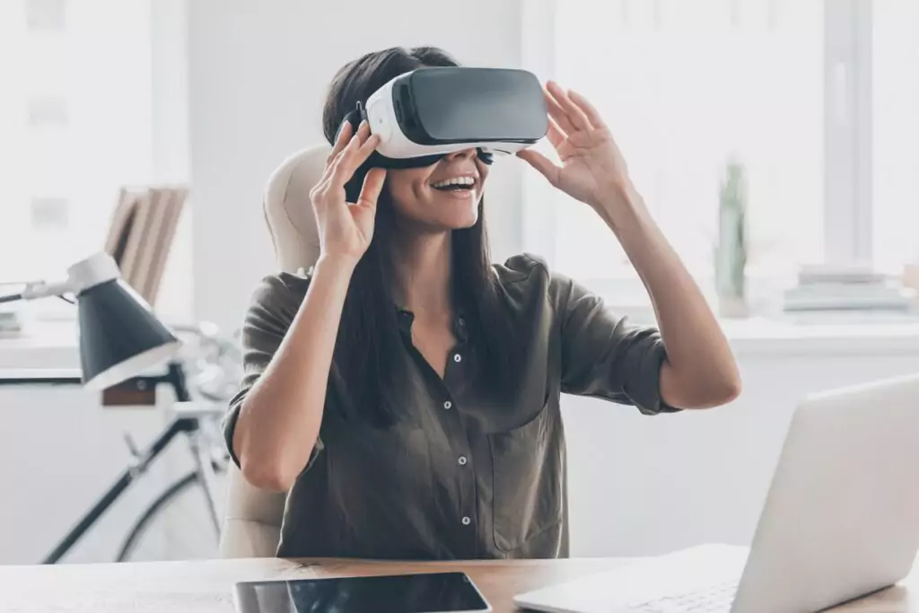 Mulher jovem confiante ajustando seu fone de ouvido de realidade virtual e sorrindo enquanto está sentado em seu local de trabalho no escritório. A realidade virtual e aumentada contribui para uma melhor experiência do cliente.
