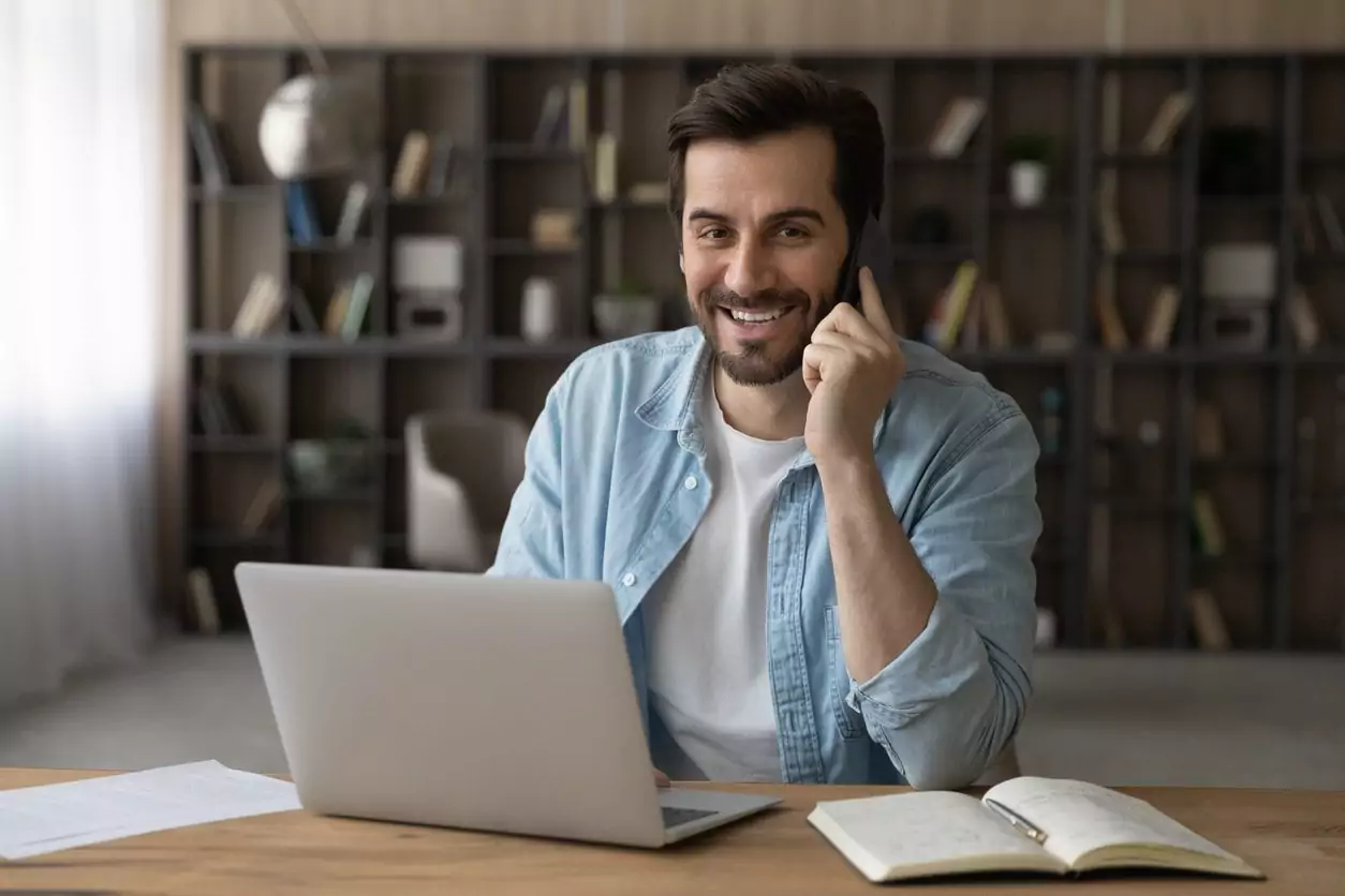Homem com jaqueta jeans sentado trabalhando com um notebook em cima da mesa e sorridente enquanto fala no celular realizando a etapa de pós-venda com seu cliente