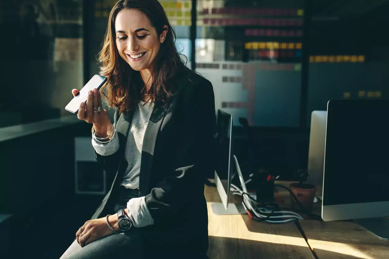 Uma mulher de cabelo curto e roupa social preta está sentada em uma mesa sorrindo enquanto segura um celular e está divulgando a sua empresa de milhas
