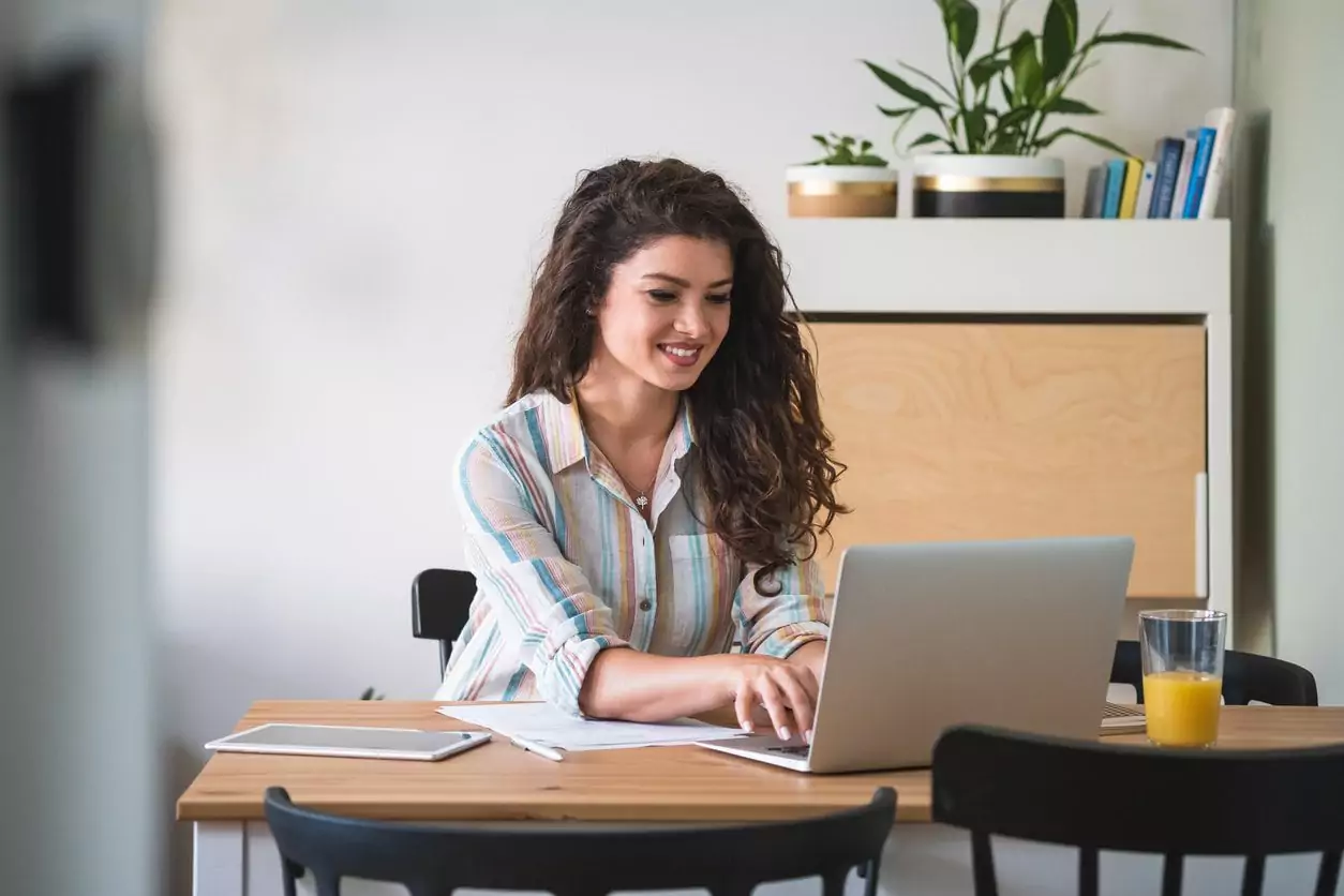 Uma mulher de cabelo ondulado e blusa listrada está sentada em uma mesa trabalhando. A moça sorri enquanto digita em seu notebook, ela está enviando um e-mail marketing para os clientes da sua empresa