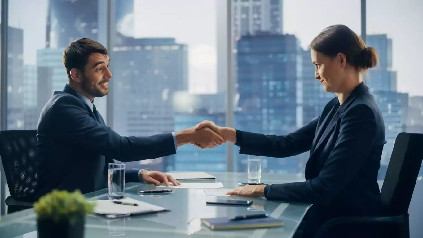 Uma mulher de coque e terno azul escuro está sorrindo e apertando a mão de um homem de terno azul escuro e cabelo preto. Eles estão selando uma parceria entre suas empresas por meio de vendas B2B realizadas entre eles