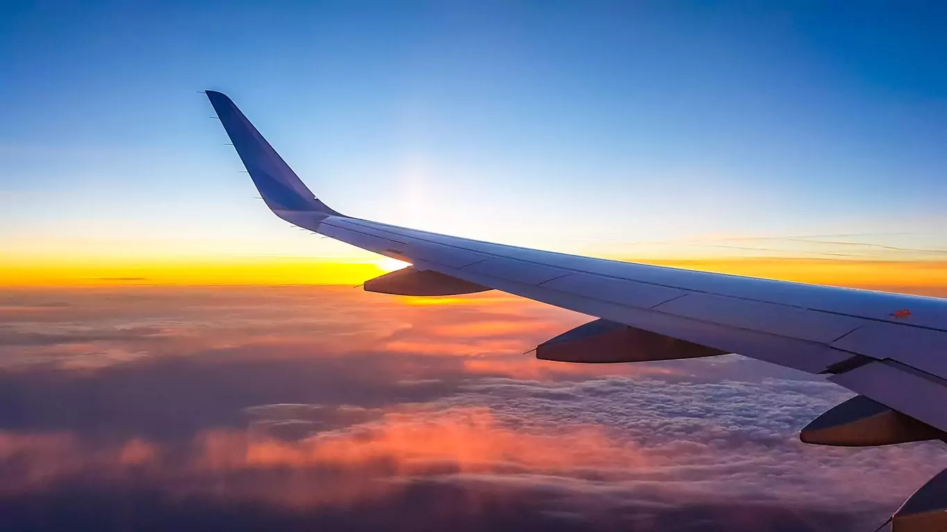 Um avião está no céu voando durante o por do sol. Os tripulantes estão com dificuldade em viajar devido ao aumento do custo do combustível de aviação