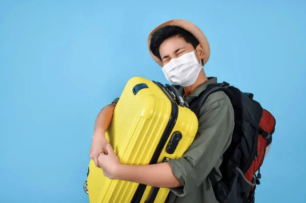 Um homem asiático,, está usando um chapéu, blusa verde e uma mochila de viagens nas costas. Ele está segurando uma mala amarela enquanto usa máscaras contra a COVID19. Ele está comemorando o boom do turismo em 2021