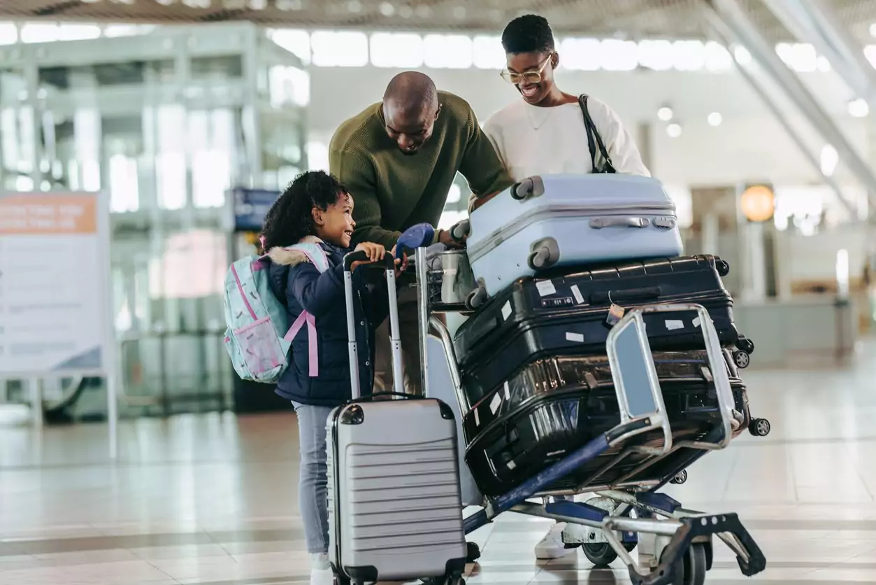 Uma família negra está empurrando um carrinho cheio de malas em um aeroporto. Eles estão sorrindo felizes porque conseguiram acompanhar as mudanças da LATAM e viajar durante as férias